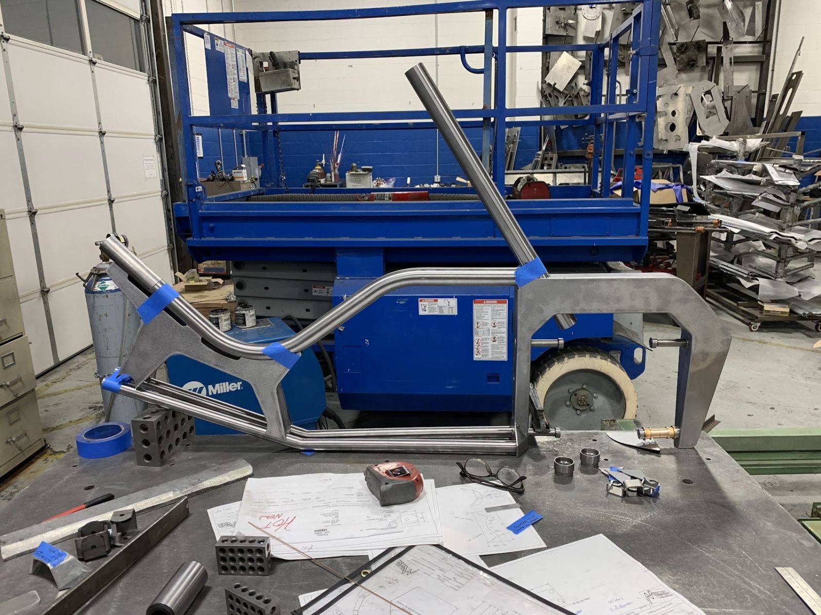 Arcimoto Mean Lean Machine frame fabrication - raw tubular suspension and steering geometry on welding table with blueprints