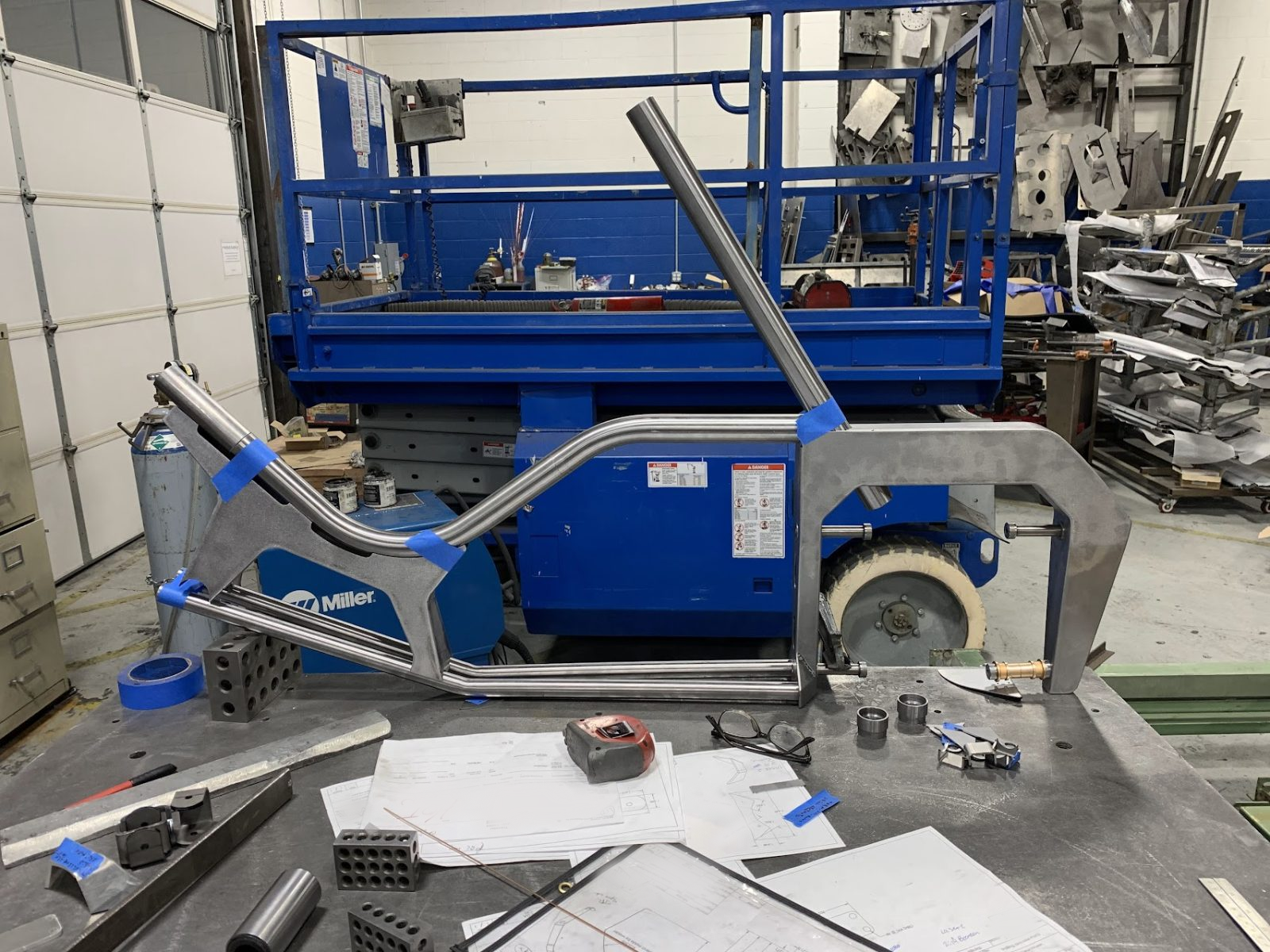 Arcimoto Mean Lean Machine frame fabrication - raw tubular suspension and steering geometry on welding table with blueprints
