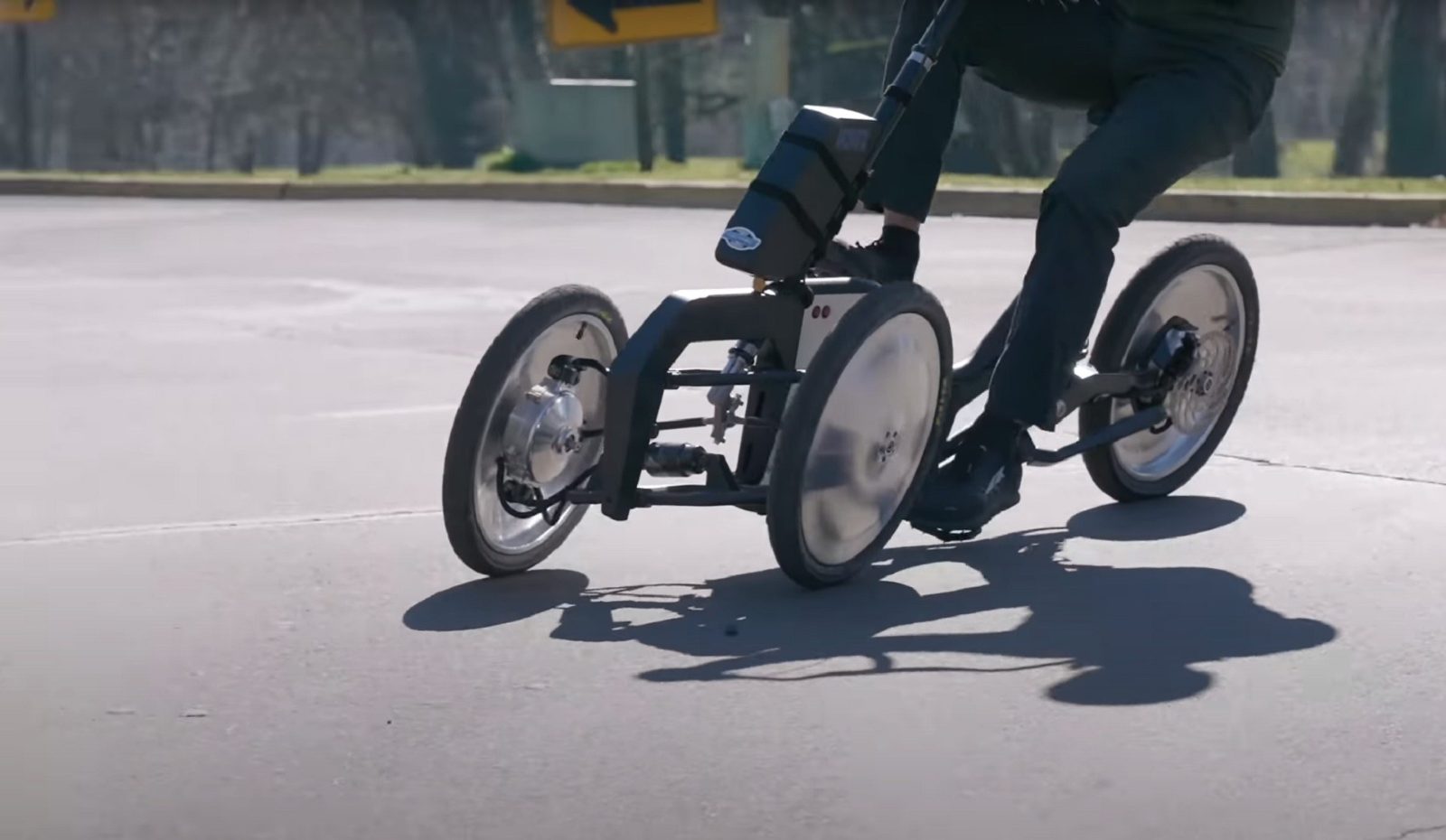 Arcimoto Mean Lean Machine in action - tilting three-wheel EV leaning through a turn on pavement, demonstrating the suspension geometry produced by the optimization framework